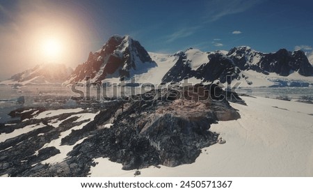 Sunset over Antarctica Mountains, Ocean. Lemaire Channel Aerial Flight. Drone Overview Of Mighty Mounts, Snow, Ice Covered Land. Bright Sun Over Ice Cold Ocean. Winter Landscape.