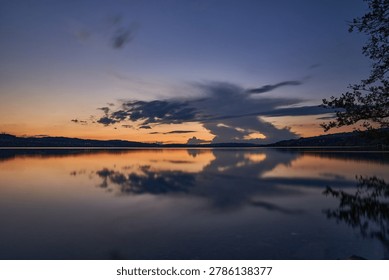 Sunset over a calm lake with dramatic cloud reflections and silhouetted shoreline