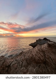 sunset over calm ocean with sharp rocky shore foreground under a dramatic sky painted with pink, orange and purple clouds