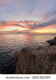 sunset over calm ocean with sharp rocky shore foreground under a dramatic sky painted with pink, orange and purple clouds