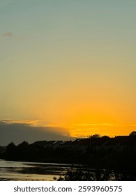 sunset over a calm river, casting a golden glow on the water. A wind turbine stands in the distance, silhouetted against the sky, while the reflection of the sun creates a peaceful atmosphere.