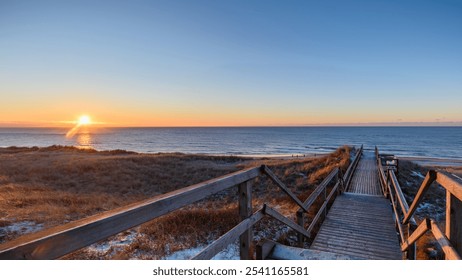 Sunset over calm sea as the golden light of sun illuminates the snowy grassy dunes and wooden pathway leading down to beach under a clear blue-orange sky. Winter on Sylt sunset over calm North Sea. - Powered by Shutterstock