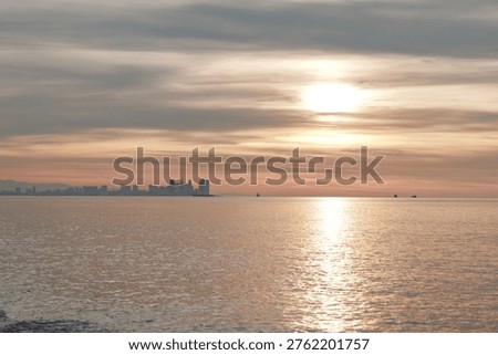 Sunset over calm water with a distant city silhouette, warm reflections on the sea and a tranquil horizon under soft clouds, creating a serene coastal scene and peaceful evening mood.