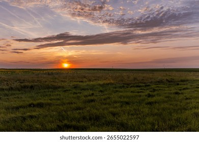 Sunset over expansive grassy fields with streaked clouds glowing in warm orange light. The sun sits low on the horizon, casting a tranquil glow across a partly cloudy sky.