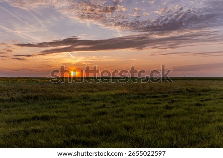 Sunset over expansive grassy fields with streaked clouds glowing in warm orange light. The sun sits low on the horizon, casting a tranquil glow across a partly cloudy sky.
