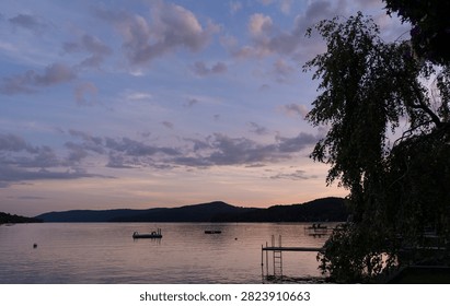 Sunset over the lake with clouds and mountain in the distance. The sun sets over the lake, casting colors in the sky as boats float on the water.