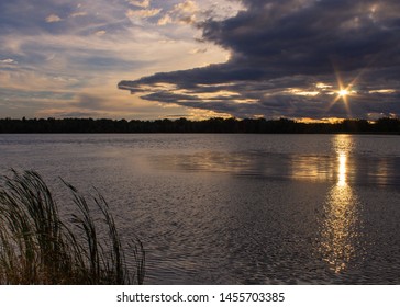 Sunset Over Lake With Sun Shining Between Clouds In Start Pattern And Reflecting On Lake. Tall Grass In Foreground. Sky Half Covered With Clounds.