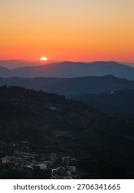 A sunset over a mountain range with a small town in the valley below. The sun is setting in the distance