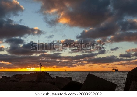 Sunset over a rocky shoreline with dramatic clouds, calm sea and a small ship on the horizon.