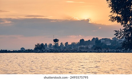 Sunset over a serene water landscape with a watchtower silhouette on the shore, framed by trees and peaceful, golden waters reflecting the warm colors of the setting sun. - Powered by Shutterstock