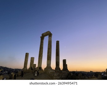 Sunset over the Temple of Hercules in Amman, Jordan. Silhouetted Roman ruins, glowing sky, crescent moon, and cityscape create a powerful, historic Middle Eastern travel scene.