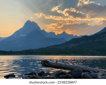 Sunset over a tranquil lake in Glacier National Park, Montana, with mountain peaks silhouetted against the vibrant sky. A driftwood log and rocky shoreline add to the peaceful scene. - Powered by Shutterstock