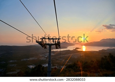 A sunset panorama featuring a central cable car tower, taut cables, the sea, mountains, and city lights below.