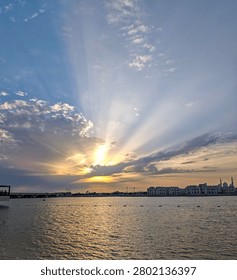 Sunset radiates golden rays over calm water. City skyline silhouettes against colorful sky