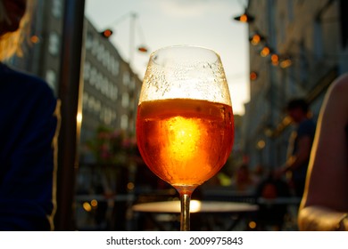 Sunset In The Restaurant Garden. On The Table A Glass Of Aperol Spritz With The Sun Breaking Through. In The Background, Buildings, Pub Lamps, The Sky. A Hot Summer Evening.