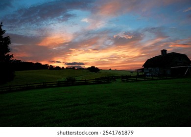 Sunset scene at a farm with barn silhouette.  Pink and blue skies with clouds.  Green pasture with wooden fence - Powered by Shutterstock