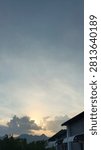 A sunset shining behind dense clouds, featuring a mountain silhouette and building rooftops in the foreground