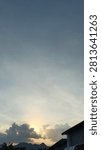 A sunset shining behind dense clouds, featuring a mountain silhouette and building rooftops in the foreground