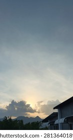 A sunset shining behind dense clouds, featuring a mountain silhouette and building rooftops in the foreground