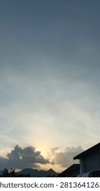 A sunset shining behind dense clouds, featuring a mountain silhouette and building rooftops in the foreground