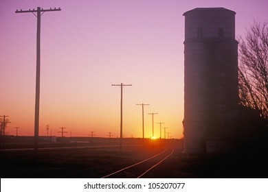 Sunset silhouette of grain silo beside train tracks, KS