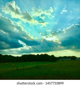 Sunset, sunrise, rays of light shining through clouds and a blue sky over agricultural farmland, hay bales, a farm, a barn and trees in rural central Virginia.