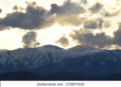 sunset or sunrise with sun glowing behind fluffy blue and white clouds over snowy mountain peaks, blue mountains, and shadowy blue foothills.  - Shutterstock ID 1728373525