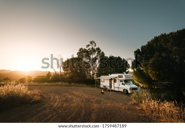 At\
sunset, a van is alone in a dirt parking lot. A fire is lit in\
front of the camper van. Sao Bartolomeu de Messines, Portugal, May\
26, 2020. Photography by Jean Baptiste\
Premat