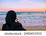 At sunset, a veiled Muslim woman stands on the beach, gazing out at the sea, her image surrounded by the golden colors of the sunset, embodying a moment of quiet contemplation.