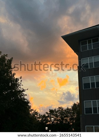Sunset view in between a campus building and trees with dark clouds overhead and a horizon filled with bright orange and yellow clouds. 