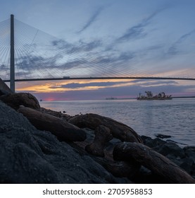 Sunset View of a Cable-Stayed Bridge with Cargo Vessel and Rusted Anchor Chain in the Foreground, Taken at the Riverbank During Blue Hour