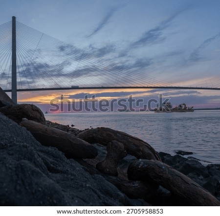 Sunset View of a Cable-Stayed Bridge with Cargo Vessel and Rusted Anchor Chain in the Foreground, Taken at the Riverbank During Blue Hour