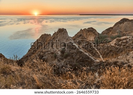 Sunset view of a coastline, with rugged cliffs and dry grass in the foreground, and the calm sea or ocean stretching to the horizon.