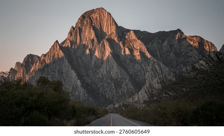 Sunset view of dramatic limestone mountains in La Huasteca Canyon, Monterrey, Mexico, with an empty road leading toward the rugged peaks under a clear sky.