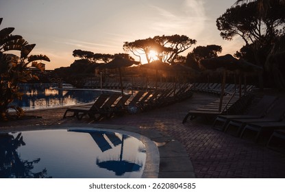 Sunset view of empty tropical resort pool area with rows of sun loungers, straw parasols, and calm water reflections surrounded by exotic trees, a peaceful vacation atmosphere; low-key wide-angle shot