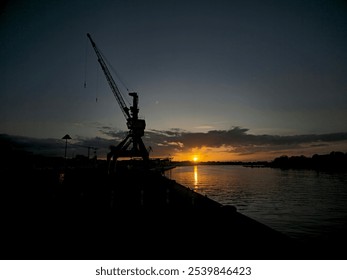 Sunset view featuring the silhouette of an industrial crane by a calm waterfront.  - Powered by Shutterstock