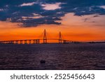 A sunset view of Incheon Bridge silhouetted against a vibrant orange and purple sky, with calm water below, showcasing the balance between nature and urban structure at Songdo Waterfront Park.