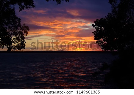 A sunset view as sun goes down behind the shade of trees over the lake. A perfect scene at the end of the day while you are relax, sitting at your patio watching a peaceful sky and birds flying home