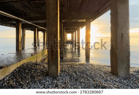 Sunset view through concrete pillars of pier over water. Beach with pebbles under pier. Calm sea reflecting golden sunlight. Pillars create leading lines toward horizon