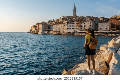 At sunset, a wanderer stands on the rocky shore, captivated by the tranquil waters of Rovinj. The historic buildings glow in the fading light, evoking a sense of adventure and peace. - Powered by Shutterstock