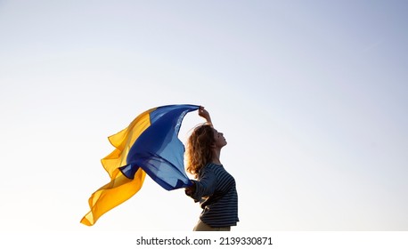 sunset, a woman's silhouette holds behind her back the Ukrainian flag fluttering in the wind. Ukrainians against the war. Drawing attention to military actions in Ukraine - Powered by Shutterstock