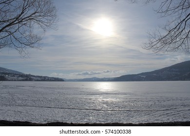 Sunshine through clouds reflecting on icy lake with tree branches in foreground. Cloudy sky, horizon and mountains in background.