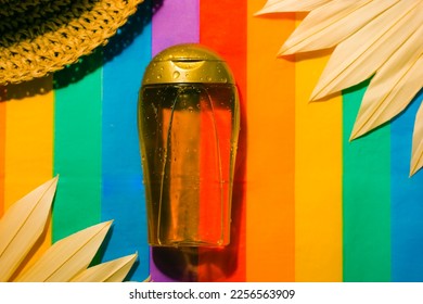 Suntan oil bottle on multicolored striped bright rainbow background. Sunblock cosmetics. Natural SPF sunscreen product for summer beach vacations. A straw hat, palm leaves. Summer cosmetic still life