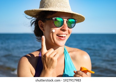 Suntan spf lotion. Beautiful woman applying sunscreen solar cream from a plastic container to her cheek, wearing straw summer hat and blue swimming suit with sea in background. Sunscreen protection.