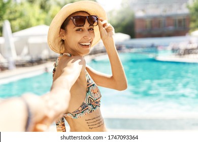 Suntanned smiling woman in hat turning around to camera and holding someone hand against swimming pool at sunny summer day. POV and Follow me concept