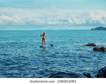 SUP Stand up paddle board. Blond girl sitting on paddle board in the sea near stone beach. Woman sailing on blue sea Mtsvane Kontskhi Beach, Batumi, Georgia.