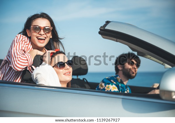 Super adventure. Portrait of happy girl in\
sunglasses is sitting in cabriolet with her friends and looking at\
camera with joy. They are travelling in car along seashore while\
enjoying summer vacation