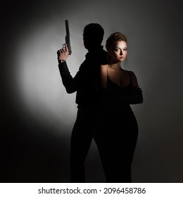 Super Agents, A Gorgeous Blonde In A Black Evening Outfit And A Man With A Gun Behind Her Back. A Luxurious Couple In Black In The Spotlight, Spy Or Noir Style