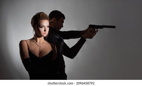 Super Agents, A Gorgeous Blonde In A Black Evening Outfit And A Man With A Gun Behind Her Back. A Luxurious Couple In Black In The Spotlight, Spy Or Noir Style