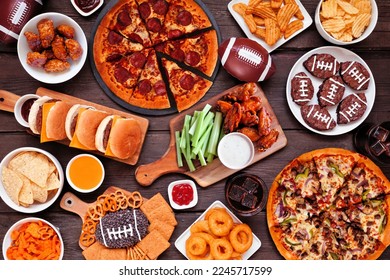 Super Bowl or football theme food table scene. Pizza, hamburgers, wings, snacks and sides. Overhead view on a dark wood background.